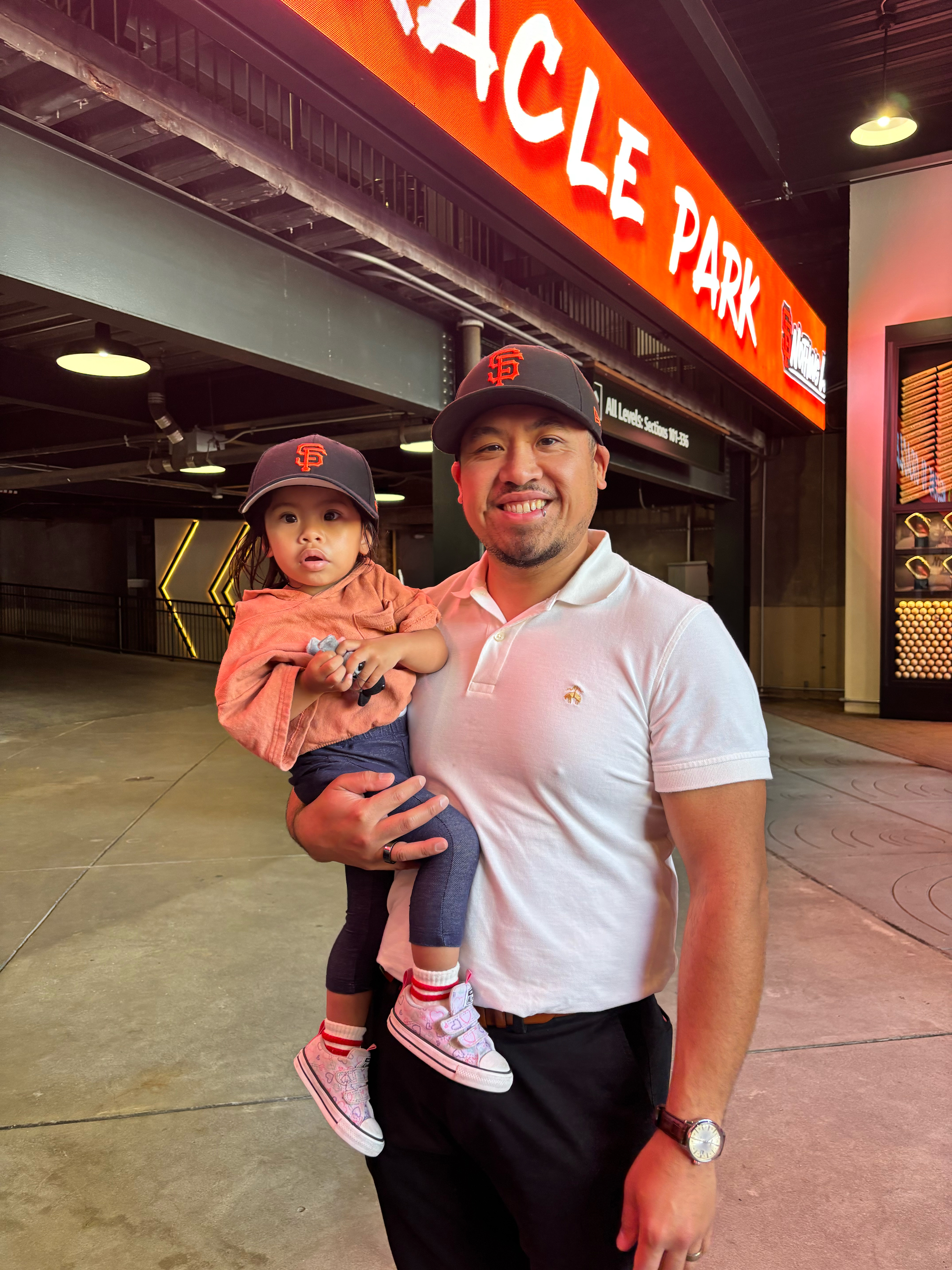 A father holds his daughter beyond the main entrance of Oracle Park, who will enjoy their first Giants baseball game together as father and daughter.
